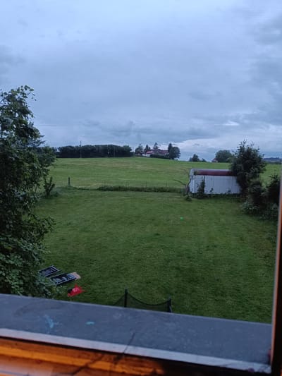 The image shows a view from a window overlooking a grassy field in Neukirch, Germany. In the foreground, visible through the window frame, are parts of a black plastic lawn mower, a red wagon, and a trampoline, partially obscured by foliage. The large green field stretches into the midground, containing a small white shed with a red roof on the right side of the frame. Beyond the field, a line of trees is visible, and in the distance, rooftops of houses dot the landscape. The sky is overcast with grey clouds. Power lines can be seen among the trees.