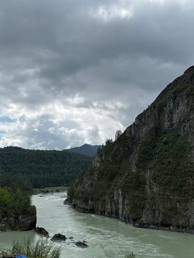 The image shows a wide, turbulent river flowing between steep, rocky mountains covered in dense green vegetation. The sky is overcast with dark, dramatic clouds. The river is a pale green color and appears to be fast-flowing, with white water rapids visible in places. The mountains on either side are rugged and imposing, with sheer cliffs and a scattering of trees. On the left side of the image, a forested hillside rises behind the river. The overall impression is one of wild, natural beauty.

This scene is consistent with the landscape found in Chemal, Russia, a popular tourist destination known for its stunning natural scenery, including rivers, mountains, and forests. The Katun River, which flows through Chemal, is a prominent feature of the area and is known for its rapids and vibrant turquoise color. The image captures the rugged, untamed beauty that draws visitors to this region.