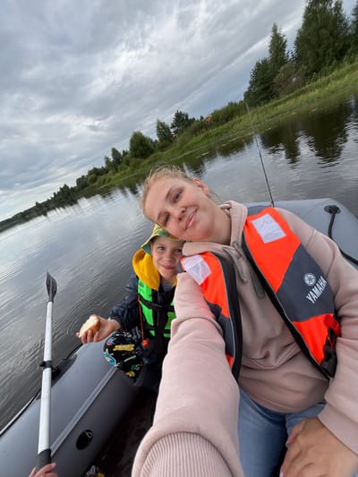 In the city of Anteplevo (Okhotinskoe S/P), Russia, a selfie is taken from a small, inflatable boat on a body of water under an overcast sky. A woman and a young boy are in the boat. The woman, in the foreground, is wearing a light pink hoodie and blue jeans, and has an orange and gray life vest on. She is smiling at the camera, with her head tilted. The boy, sitting behind her, is wearing a yellow hat and a green and black life vest. He is holding a piece of bread. A paddle is visible on the left side of the boat, and a fishing rod is propped up on the right. The background shows a tree-lined shore with some buildings in the distance. The water is calm with ripples reflecting the cloudy sky.