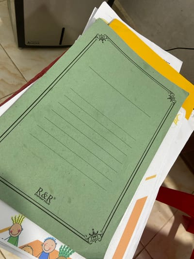 The image shows a stack of papers and folders. A green folder with black decorative borders and horizontal lines is prominently displayed. Text on the green folder reads "R&R STOPMAP FOLIO". Below the green folder, a white paper with cartoon drawings of children is visible. A yellow folder is tucked behind the green folder. The background shows a tiled floor and a part of a dark gray cabinet with the word "ALAIXA" visible on it. A red object, possibly a chair, is partially visible on the right side of the image. The scene appears to be indoors, likely in an office or study area. No specific location in Tanjung Gading, Indonesia can be identified from the image.