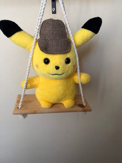 A plush Pikachu toy wearing a brown herringbone cap is seated on a small wooden swing suspended by white ropes against a beige, textured wall. The toy is yellow with black tips on its ears and has red blush marks on its cheeks. The swing is made of light-colored wood and has two holes through which the ropes pass. The ropes are attached to an unseen point above the toy. The lighting is even, casting soft shadows. The scene appears to be indoors, possibly a domestic setting. There is no indication of the location being Dzhalgan, Russia.
