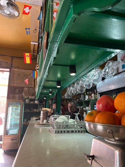 The image shows the interior of a bar, likely a tapas bar, in Agüimes, Spain. It is situated at a location defined by the coordinates 27.907 latitude and -15.447 longitude. The bar is decorated with a green ceiling and wooden beams, and there are several flags hanging from the ceiling. The bar is filled with a selection of wine bottles on the back shelf, empty glasses hanging from the ceiling, and various plates and utensils stacked on the counter. A few people are present: a man in a white shirt with a ponytail, a man behind the counter with a black shirt and short hair, and another man sitting at a table on the left-hand side of the image. There is a scale and a bowl full of oranges and apples in the foreground.