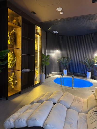 The image depicts a dimly lit spa room, likely a private wellness area. The room features a small, circular pool with vibrant blue water, accessible by a short set of chrome railings. Two potted palm-like plants stand on either side of the pool. The walls are decorated with dark, vertical slatted panels, and a dark, circular acoustic ceiling feature is visible.
To the left of the pool, a tall, dark wooden shelving unit is illuminated by warm, vertical strip lighting. Several shelves display dried, yellowed bundles, possibly of herbs or branches, reminiscent of bathhouse whisks, along with some decorative yellow objects. One shelf in the center has a folded white cloth.
In the foreground, a series of large, white, rectangular floor cushions are arranged in rows, suggesting a relaxation area. The floor is tiled with a light-colored stone. The overall atmosphere is serene and luxurious.
The specific location in Chingeldi, Uzbekistan, cannot be determined from the visual information provided in the image.