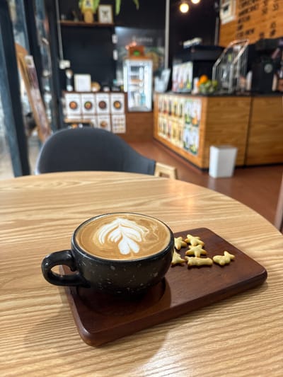 A close-up shot shows a dark speckled mug of latte art coffee and a small wooden tray with several star-shaped crackers on a light-colored wooden table. The mug is black with a white latte art design, resembling a leaf. The wooden tray is dark brown and rectangular with rounded corners. The crackers are light golden brown and appear to be sweet biscuits. In the blurred background, a cafe interior is visible, with shelves displaying items and a counter with some bottles and fruits. This scene is captured within a cafe located in Tha Chana, Thailand.