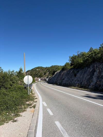 The image shows a road in Kućišta, Montenegro. The road is asphalt with white dividing lines and curves to the right. On the left side of the road, there is a gravel shoulder with a metal guardrail, a concrete pole, and a circular white sign. Dense green vegetation grows behind the guardrail. To the right of the road, a steep rocky cliff face rises, covered in patches of green vegetation. In the distance, a tunnel entrance is visible on the mountain. The sky is a clear, bright blue. There are no people or vehicles visible in the image.