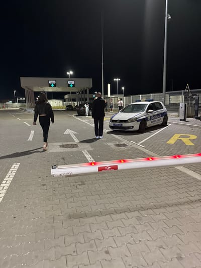 The image depicts a border crossing point at night, situated in Prijepolje Municipality, Serbia. A white border police vehicle is parked in a reserved parking area. Two people, a man and a woman, are walking towards the border control building. A traffic barrier stands near the pedestrian walkway. The ground is paved with interlocking bricks. Streetlights illuminate the area, and border control signs are visible on the building.