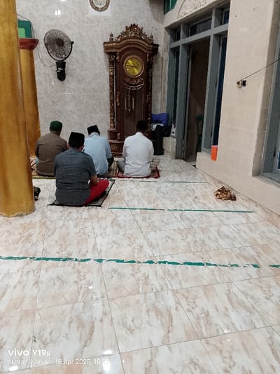 The image shows a mosque interior in Kecamatan Kroya, Indonesia, located at coordinates -6.423 latitude and 107.996 longitude. The scene depicts four adult males engaged in prayer, kneeling on prayer rugs, facing a large wooden clock. The clock stands against the wall, with a Roman numeral clock face, a wooden pendulum, and a gold-painted top. The walls are tiled in white, and the floor is tiled in a beige and brown pattern. The men are wearing traditional Islamic clothing, and their prayer rugs are red and green. There is a large window in the back, with a door leading out.
