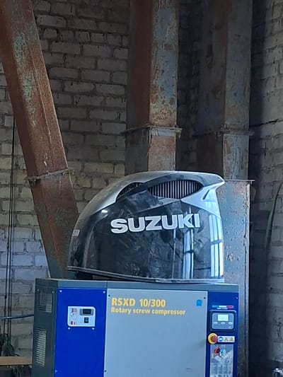 This is a medium-shot, eye-level, slightly angled photograph taken outdoors in Žemaitkiemis, Lithuania. The scene depicts a rotary screw compressor, likely industrial equipment, situated in what appears to be an outdoor, possibly agricultural or workshop setting. The compressor is primarily colored blue and gray, with a prominent black cover on top bearing the white "SUZUKI" logo and model designation "RXSD 10/300 Rotary screw compressor". Behind the compressor, a brick wall and large, rusty metal pillars are visible. The overall impression is one of an industrial or functional space, with weathered structures and equipment. There are no people or animals present in the image.