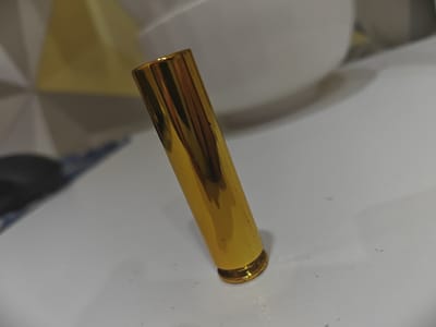 The image shows a close-up, low-angle shot of a single, metallic gold, cylindrical object, possibly a lipstick tube or a pen cap, standing upright on a white, textured surface. The object is positioned slightly off-center, towards the right side of the frame, and is angled to the left. Its surface is highly reflective, showing streaks of light and darker areas that suggest the surrounding environment. The background is out of focus, with hints of a white bowl-like object and a yellow geometric pattern. There is no indication of the object's location within Bugulma, Russia, or any activity related to it.