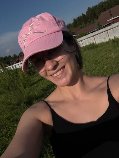 A selfie is taken from a low angle outside on a sunny day, with a young woman wearing a pink baseball cap with a white bow embroidered on it and glasses. She has a black tank top on and is smiling. In the background, there is a white metal fence, some trees, and houses with brown roofs. The ground is covered in green grass and some small pine trees. The sky is blue with a few clouds. The location is identified as Doksha, Russia.