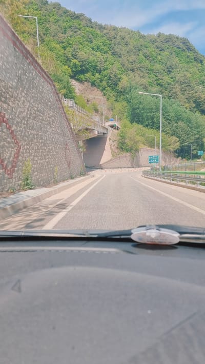 The image depicts a view from a vehicle, possibly a bus, driving along a road in Yongpyeong-myeon, South Korea. The road is bordered by a stone retaining wall on the left and a mountain covered in trees on the other side. A pedestrian tunnel entrance is visible on the mountain side, accessible by a set of stairs. Roadside signage and lighting poles are present along the road. Part of the vehicle's interior is visible in the foreground, including a portion of the dashboard and a circular object attached to the windshield. The sky is visible in the upper left corner.