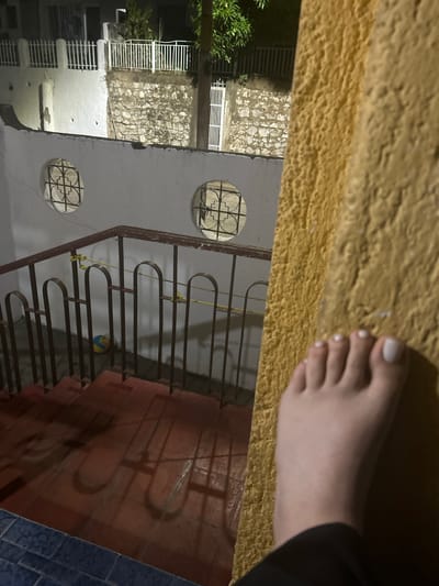 A close-up shot focuses on a person's foot with white nail polish on a rough, yellow surface. In the background, through a railing, is a view of a building. The building features decorative round windows and a wall with stone or brickwork. A ball is visible on the ground level of the building. The scene suggests a casual, possibly nighttime or low-light setting. There is no verifiable evidence in the image to support that it was taken in San Juan de Los Morros, Venezuela.