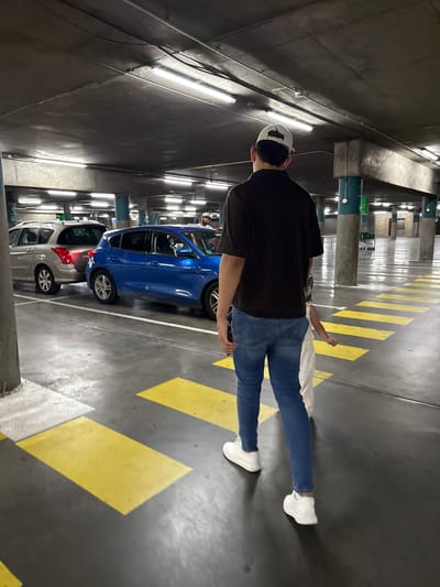 In a multi-level concrete parking garage, a man wearing a black t-shirt, blue jeans, white sneakers, and a light-colored baseball cap walks away from the camera, followed by a young child in a white dress. The man appears to be holding the child's hand. They are walking on a dark gray concrete floor marked with yellow painted rectangles. Several parked cars are visible in the background, including a silver station wagon and a bright blue hatchback. The garage is illuminated by fluorescent strip lighting. The environment is consistent with a city parking structure, potentially in an urban or suburban area. The presence of cars and the architecture suggest a location where people park their vehicles, such as a shopping center, office building, or residential complex. Without further information, it is impossible to definitively place this scene in Carretera De San Martín, Spain.