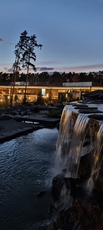 The image shows an outdoor scene at dusk with a modern building in the background. The building has large windows with interior lights illuminating the interior spaces, suggesting it is a place of business or residence. Trees are visible surrounding the building, with a few tall pine trees in silhouette against the twilight sky on the left.

In the foreground, there is a man-made waterfall cascading over rocks into a body of water. The water is illuminated by the artificial lights from the building, creating a reflective surface. The sky is a gradient of blues and soft pinks, typical of sunset or twilight. The overall mood is peaceful and serene, with the gentle sound of the waterfall implied. There is no visible text in the image. The location context of Shulgino, Russia, suggests a possibly tranquil, perhaps suburban or park-like setting. The image captures a moment of transition between day and night, highlighting the interplay of natural and artificial light.