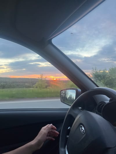 The image is a first-person perspective from inside a car, looking out the windshield and side window towards a sunset. The sky is filled with dramatic clouds illuminated by the setting sun, casting an orange glow over the landscape. The landscape appears rural, with a line of trees in the distance and a field of green grass in the foreground, bordering a road. The interior of the car is visible, including a steering wheel and a hand resting on the door panel. A side-view mirror reflects the trees and sky. There is no visible information to determine the specific location within Забегалово, Russia.