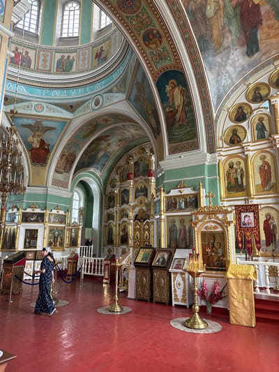 This image shows the interior of the Church of the Resurrection of Christ in Nogornyi, Russia. A woman in a long, dark floral dress and a headscarf walks across a bright red floor. The church's interior is elaborately decorated with golden icons and religious frescoes adorning the walls and domed ceiling. A large, ornate iconostasis dominates the right side of the frame, featuring numerous paintings of saints and religious scenes. A tall, golden candelabra with lit candles stands in the foreground. The overall impression is one of grandeur and religious devotion.