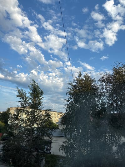 The image depicts a view from a window, looking upwards towards a bright blue sky filled with scattered white, puffy clouds. In the foreground, the dark silhouettes of trees with dense foliage obscure part of the view. Behind the trees, several multi-story apartment buildings with light-colored facades are visible. A thin black wire stretches diagonally across the upper portion of the image. The scene appears to be set in the city of Lysva, Russia, as indicated by the architecture and general urban landscape. There are no people or observable actions taking place.