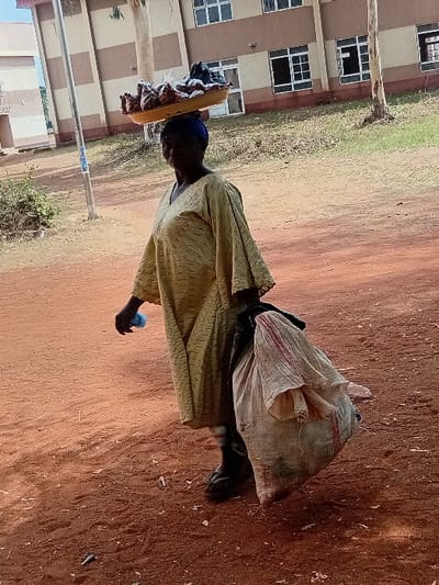 An outdoor image, rotated 90 degrees clockwise, depicts a person walking or standing on a reddish-brown dirt path in front of a building. The individual is seen from the side, facing left (when oriented upright). They are wearing a light-colored, possibly patterned, long-sleeved garment and a dark blue head covering.
On their head, they are balancing a flat, rectangular, light-colored tray or board, on which several dark-colored items, possibly wrapped food portions, are placed. In their left hand, they hold a small, blue, somewhat fluffy object.
To the person's right, resting on the ground, is a large, light-colored, possibly woven sack or bag, which appears full. The ground beneath them is predominantly exposed reddish-brown earth, with some patches of green grass visible further back.
In the background, a multi-story building with a light brown or tan facade is visible. It features numerous rectangular windows, many of which have white frames and multiple panes. A slender, light-colored pole or tree trunk stands vertically in the mid-ground, slightly to the left of the person.
The scene is well-lit, suggesting daytime under clear skies. No text is visible in the image.