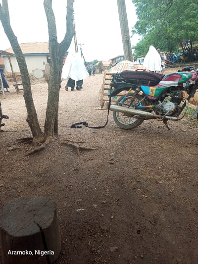 The image shows a motorcycle parked in front of a tree trunk, with a black strap attached to it, located in Aramoko, Nigeria, at coordinates (latitude: 7.835, longitude: 4.479). The motorcycle is red, green, and blue, with a black seat and a black exhaust pipe. The tree trunk is brown and has visible roots. In the background, a group of people are walking away from the camera, all wearing white clothing. The ground is dirt and gravel. There is a wooden log on the ground in the foreground. The scene appears to be a street in a town or village, with a dirt path leading away from the parked motorcycle.