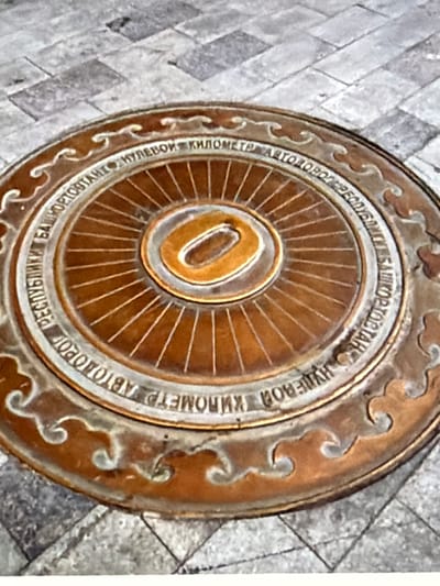 A close-up, overhead view captures a circular bronze plaque embedded in a cobblestone surface. The plaque is adorned with raised lettering that encircles a central design. The text, appearing to be in Russian, reads "КИЛОМЕТР АВТОДОРОГ РЕСПУБЛИКИ БАШКОРТОСТАН" which translates to "Kilometer of the Roads of the Republic of Bashkortostan." The central design features radiating lines emanating from a hollow oval shape in the very center, all rendered in bronze with a golden hue. The plaque is set into a ground composed of uniform, grey, rectangular cobblestones arranged in a diagonal pattern. The overall image presents a static, outdoor scene of a landmark.
