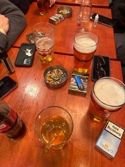The image displays a reddish-brown wooden table in an indoor public establishment located in Vincennes, France. The scene is presented from an overhead perspective, focusing on various items associated with a social gathering involving multiple individuals.
**Objects and Subjects:**
* **Beverages:** Four glasses containing amber liquid are present. Three are pint glasses, approximately half to three-quarters full, each topped with a white foam head, indicative of beer. The fourth is a clear glass tumbler containing amber liquid with a clear glass straw. A partially consumed brown glass bottle with a red label, identifiable as "Coca-Cola zero sucre," is also on the table. An empty stemmed wine glass stands in the upper right section.
* **Smoking Paraphernalia:** Two small, circular metallic ashtrays are visible. One is filled with numerous cigarette butts and ash. The other, in the upper section, contains fewer butts. Three cigarette packs are present: one largely obscured, another partially wrapped in plastic with a blue Bic lighter resting on top, and an unopened "Camel Blue" pack oriented upside down, displaying the English health warning "Smoking Kills."
* **Electronic Devices:** A black smartphone encased in a black cover featuring a white graphic design and dual camera lenses is centrally located. Another black smartphone without a case is visible in the lower left. A black vape pen or e-cigarette device stands upright on the left side.
* **Personal Items:** A pair of black-framed eyeglasses is positioned in the middle right section. A small white plastic dropper bottle is near the empty wine glass.
* **Human Elements:** A portion of a person's left arm, clad in a grey long-sleeved garment, is visible in the upper left. A white wristband displaying "PARIS 2024" is on this arm. A dark fabric, likely part of another person's clothing, is partially visible in the upper right.
**Actions and Interactions:**
The collective presence of multiple drinks, full and used ashtrays, and various personal items indicates that several individuals are actively consuming beverages and tobacco products in a social setting. No explicit actions are depicted, but the arrangement implies ongoing interaction and leisure.