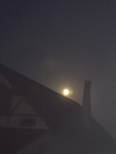 The image shows a dark, misty night sky over a silhouetted roofline in Kostelec na Hané, Czechia. The moon is visible as a bright, hazy orb partially obscured by the mist, casting a soft glow. A chimney stands prominently on the right side of the frame, also silhouetted against the sky. The roof appears to be part of a residential building, with subtle architectural details suggested by the dark shapes. The overall atmosphere is moody and ethereal, with the fog softening the edges of the structures and enhancing the luminescence of the moon.