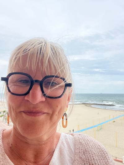 The image shows a woman with short, blonde hair and glasses standing on a balcony overlooking a beach in Palavas-les-Flots, France. She wears a white top and a light pink cardigan. The beach below has a blue barrier with yellow posts. The sky is cloudy, and the sea is visible in the background.