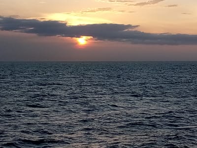 The image shows a sunset over the ocean. The sun is partially obscured by clouds, casting a warm glow across the sky. The ocean is dark and choppy, with small waves. The horizon is visible, separating the sea from the sky. The location is not specified in the image, therefore it cannot be identified as being in the city of Unknown, Venezuela.
