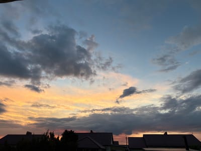 This is a landscape photograph taken at dusk in Uzylukovo, Russia. The image features a dramatic sunset sky filled with a mix of dark, heavy clouds and streaks of vibrant orange, yellow, and pink. These colorful clouds suggest the sun is low on the horizon, casting a warm glow across the sky.

Below the sky, silhouetted against the fading light, are the rooftops of several houses, indicating a residential setting. Dark green trees are also visible in the foreground, adding to the silhouette effect. The roofs are dark, and some have chimneys. There are also power lines visible stretching across the scene.

The overall mood of the image is serene and contemplative, characteristic of a sunset. The weather appears to be partly cloudy, with the clouds creating an interesting visual texture and contrast with the colorful sky. The location is identified as Uzylukovo, Russia, adding a geographical context to the scene. There is no visible text in the image.