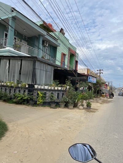 The image shows a street scene in Landuh, Indonesia, likely in the morning or early afternoon due to the bright but somewhat overcast sky. The perspective is from someone on a motorbike, looking towards a multi-story building on the left and the road on the right.
The building is painted in shades of light green and teal, with orange accents around some windows. It has multiple levels, with balconies and windows visible. There are many power lines crisscrossing the scene above the building and road. Along the base of the building, a row of potted plants is displayed, creating a green border. Some of these plants are lush and full, while others are smaller.
The road appears to be a dirt or gravel road, with some patches of wetness or mud. To the right of the plants, the road extends into the distance, where other buildings and vehicles can be seen. There's a street sign visible with text that is difficult to read. A banner is also hanging from a pole, with the word "PRESTIGE" clearly visible in large blue letters.
In the reflection of the motorbike's rearview mirror, a person wearing a light blue garment is visible, indicating the photographer's presence. The overall scene suggests a typical street in a developing area, with a mix of residential or commercial buildings, natural elements like plants, and the ubiquitous presence of overhead electrical wiring. The weather appears to be partly cloudy, with the sun occasionally breaking through.