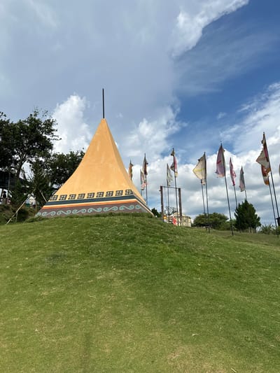 The image depicts an outdoor scene on a grassy hill, likely in the city of Thôn 5, Vietnam, given the context. The central focus is a large, orange, pyramid-shaped structure with decorative bands around its base, featuring geometric patterns and wavy designs. A thin, black pole extends vertically from the apex of the structure. To the right of the pyramid, a line of tall flagpoles displays various colorful flags, many appearing tattered or wind-worn. In the background, trees are visible, and a swing set with a wooden frame is present. The sky above is partly cloudy with patches of blue sky. The ground is covered in short, green grass. There are no visible people directly interacting with the central structure or flags in the foreground.