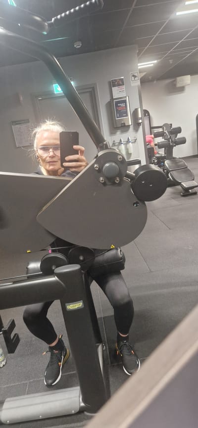 The image shows a woman in a gym, taking a selfie in a mirror. She is wearing glasses, black leggings, and black sneakers with orange accents. She is sitting on a piece of gym equipment that appears to be a leg press machine. The gym environment is visible in the background, with other weight machines and a water bottle on the floor. The image does not contain any information that would indicate the location of Stockholm, Sweden.