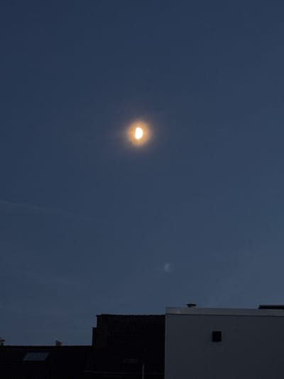The image shows a sliver of the moon in a dark blue sky, above several buildings. The moon is illuminated on its right side, with the left side in shadow. One building in the foreground is dark and appears to be a residential building with a skylight. Another building to the right is white with a dark square window. The sky is clear with no clouds visible. The location is Molenbeek-Saint-Jean - Sint-Jans-Molenbeek, Belgium.