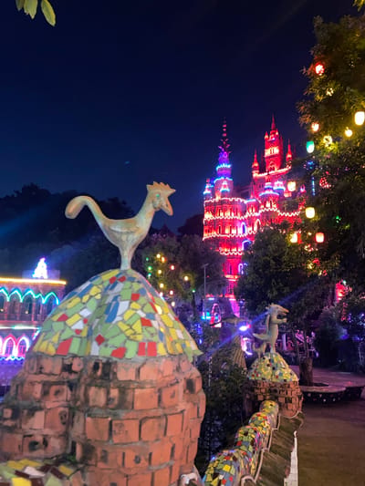 This image depicts the night view of the Baqiao Baqiao Scenic Area in Baqiao, China. In the foreground, a mosaic-covered brick structure supports a decorative ceramic rooster sculpture. To the right, another smaller mosaic-covered brick structure is topped with a ceramic dragon sculpture. In the background, a large, ornate building is illuminated with vibrant red and blue lights, resembling a castle. A tree decorated with colorful lanterns stands to the right of the building. The sky is dark blue, indicating it is nighttime. The scene is decorated with numerous colorful lights, creating a festive and fantastical atmosphere.