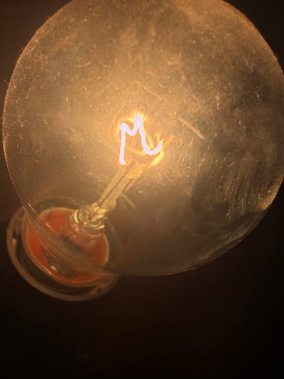 An extreme close-up shot captures the filament of an illuminated light bulb. The filament, glowing brightly with a bluish-white light, forms the shape of the letter "M". It is suspended within a clear glass bulb that appears dusty and slightly smudged, creating a soft, diffused illumination. The base of the bulb, where it connects to a socket, is visible at the bottom and appears to be a dark, reddish-brown material. The background is a deep, uniform black, indicating a lack of ambient light in the environment. This image provides a forensic, matter-of-fact depiction of the internal structure and illumination of a light bulb, with no humans or specific environmental context beyond what is intrinsic to the object itself. The location is not discernible from the image.