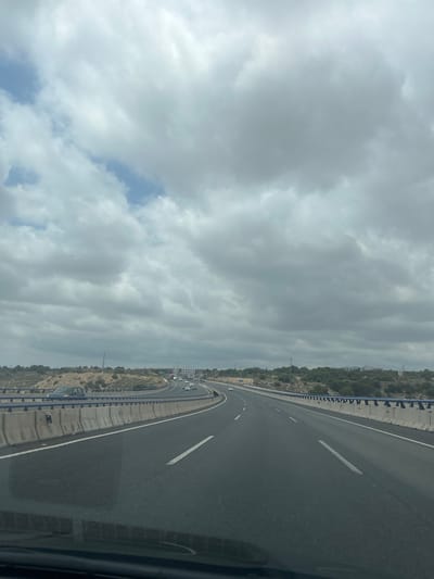 The image shows a view from a moving vehicle on a multi-lane highway. The highway curves to the right in the distance. The sky is heavily overcast with thick, gray clouds. The landscape on either side of the highway consists of dry, arid terrain with sparse vegetation and some hills. Concrete barriers and metal railings line the sides of the road. Several cars are visible on the highway, driving in the same direction as the viewpoint. The overall scene suggests a drive through a somewhat desolate, possibly rural or suburban, area under a cloudy sky. The provided information suggests the location is the city of Elx / Elche, Spain.