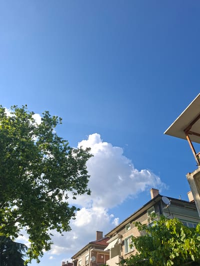 In the city of Aksakovo, Bulgaria, this image captures a clear blue sky with scattered white clouds. On the left, a large green tree with dense foliage dominates the foreground. To the right, the upper portions of buildings are visible. The closest building on the right features a pale yellow and grey facade with multiple windows. A satellite dish is mounted on its roof, which also has a brick chimney. Above this building, a white canopy extends from a concrete structure. Further to the left and behind the main building, another building with a red-tiled roof is partially visible. The overall scene depicts an urban environment under a bright sky, with natural elements like the tree contrasting with the man-made structures.