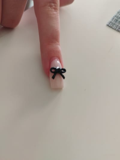 A close-up, overhead shot shows a person's left index finger against a pale, light-colored surface. The finger has a light pink, polished nail with a small, black bow charm attached to the tip. The bow is centered on the nail and appears to be made of a flexible material. The finger itself is smooth with visible skin texture. The background surface is uniform and appears to be a table or desk. To the upper right of the finger, a small, gray, textured object, possibly fabric or a textured sticker, is visible. The lighting is even, suggesting an indoor environment, and no specific location in Meyrin, Switzerland can be determined from the image.