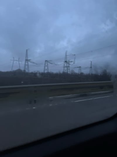 A gray, overcast sky looms over a line of electrical towers extending into the distance. The structures are thin and metallic, carrying power lines across a landscape blurred by rain or fog. Dense trees fill the space between the towers. The photograph appears to have been taken from inside a vehicle, as a portion of the window frame is visible on the right side, with water droplets adding to the indistinctness.