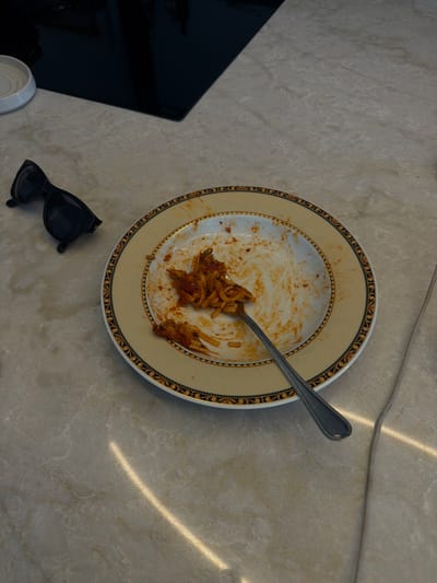 A close-up, high-angle shot shows a nearly empty plate of pasta with red sauce and a fork resting on a marble-patterned countertop. To the left of the plate, a pair of black sunglasses are laid down. In the background, a black glass stovetop is visible, with a white circular ceramic dish to its left. The scene is located in Ridderkerk, Netherlands.