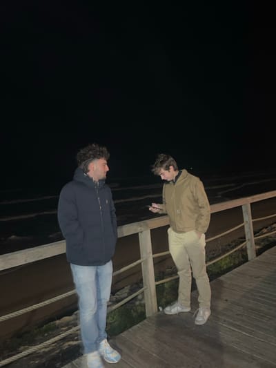 Two young men stand facing each other on a wooden walkway at night. One wears a tan jacket and light-colored pants, looking down. The other wears a dark blue puffer jacket and jeans, gazing upwards. The walkway has a simple wooden railing with ropes. Beyond the walkway is a dark, undefined area. The dark sky occupies a significant portion of the upper left frame.