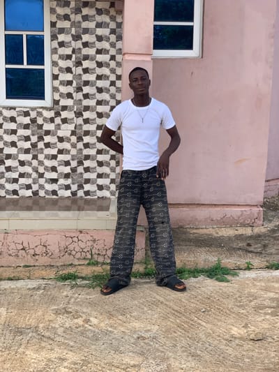 A young man stands in front of a building. He is wearing a white T-shirt, black pants with a repeated pattern, and black sandals. His hands are on his hips. Behind him is a tiled wall, a light pink wall, and a concrete walkway with patches of vegetation. A window is visible on the left side of the frame. The photo was taken in Gbeleko, Nigeria.