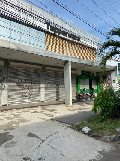 The image shows the exterior of a closed storefront with a sign that reads "Tupperware" in Purwakarta, Indonesia. The storefront has three roll-down metal doors that are closed. The doors are marked with graffiti. There are two motorcycles parked in front of the storefront. The scene is taken from a street level view. There are multiple power lines running across the top of the building. There is a green-colored storefront to the right of the Tupperware storefront. There is also a small palm tree to the right of the Tupperware storefront and a small bush to the left of the green storefront. There are no people visible in the image.