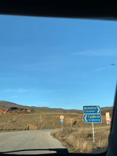 The image, taken from inside a vehicle looking through its windshield, captures a sunny daytime scene with a vast, clear blue sky overhead. The perspective is slightly rotated clockwise, showing the horizon line diagonally across the frame.

The central focus of the image is on two prominent blue directional road signs with white text, indicating destinations. The sign on the left displays "Δεσκάτη" (Deskati) in Greek and "Deskati" in English, suggesting a route forward or slightly to the right. The sign on the right, pointing more sharply to the right, reads "Γρεβενά" (Grevena) in Greek and "Grevena" in English. These signs are typical of road signage in Greece.

Below and to the right of these blue signs, a standard red and white international STOP sign is clearly visible. Adjacent to the stop sign is a circular blue mandatory instruction sign featuring a white arrow pointing to the left, indicating a required turn or direction.

The landscape beyond the road signs consists of dry, grassy hills and sparse vegetation, consistent with a rural or semi-arid environment. The road itself appears to be asphalt, curving off to the right. The bright, cloudless sky indicates clear weather conditions. The edges of the car's interior, likely the dashboard or windshield frame, are visible on the left and right sides of the image. The presence of the Greek town names "Deskati" and "Grevena" explicitly confirms the location as Greece, with the user context placing it around Grevena.