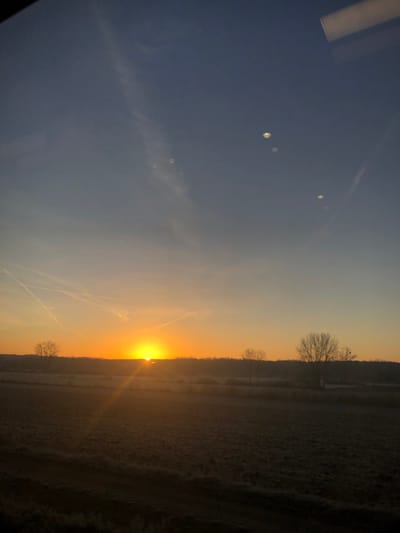 A vibrant sunrise illuminates a frosty landscape, transitioning from a dark foreground to a bright horizon. The sky fades from deep blue to golden orange. Silhouetted trees line the horizon, adding depth to the scene. Vapor trails crisscross the sky, adding linear details. The sun is a central focal point, radiating light and warmth. Reflections or light flares appear in the upper left corner.