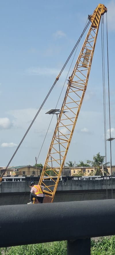 The image shows a yellow lattice boom crane against a partly cloudy sky, likely in an urban area. The crane is angled upward and leftward. A worker wearing a safety vest and helmet is visible near the base of the boom. Buildings with palm trees are visible behind the crane. A black cylindrical structure (possibly a pipe or pillar) is in the foreground, partially obscuring the view. Based on the location information, this scene is likely in the city of Satellite Town, Nigeria.