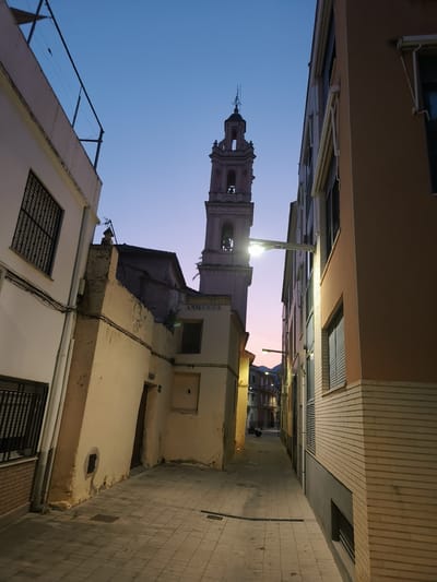 The image depicts a narrow street in Benirredrà, Spain, at dusk. The sky is a gradient of deep blue at the top transitioning to pale pink and orange near the horizon. Dominating the scene is a tall, ornate bell tower, likely part of a church, with architectural detailing and a bell visible in one of its openings. The tower is made of a light-colored stone, possibly pinkish hue, and is illuminated by artificial light.

Flanking the street are buildings on either side. The building on the left is lighter in color with windows featuring dark metal grates. The building on the right is a warm peach color with brick detailing on the lower portion. This building has multiple windows, some with horizontal blinds, and external light fixtures are mounted on its facade.

The street itself is paved with light-colored square tiles and features drainage grates running along its sides. The perspective suggests the camera is looking down the street, away from the viewer, towards the distant buildings and the colorful sky. There are no people visible in the image, and the scene appears quiet and serene. The overall impression is one of a charming European street bathed in the soft light of twilight. The date "ANO 1892" is visible on the side of a building closer to the tower.