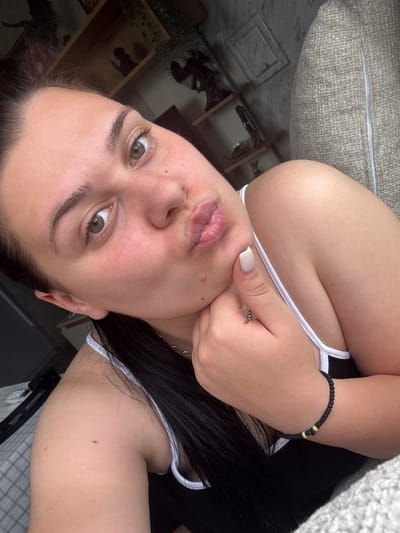 A young woman is captured in a close-up, vertically oriented selfie. She is wearing a black tank top with white trim. Her right hand is near her face, with her thumb gently touching her cheek, and she is wearing a ring on her index finger. Her lips are pursed, and she has light-colored eyeshadow on. She also wears a dark braided bracelet with two small neon green beads on her left wrist. The background shows a textured, light-colored couch or cushion, and hints of furniture and possibly a shelf with items in the lower left quadrant. The lighting suggests it is daytime. The woman's expression is playful or inquisitive.