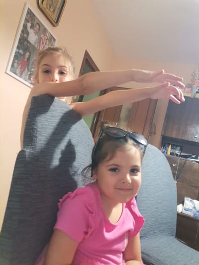 The image shows two young girls in what appears to be a home setting. The girl in the foreground is wearing a pink shirt with ruffled sleeves and has sunglasses perched on her head. She is smiling at the camera. Behind her, another girl, with blonde hair, is partially visible, resting her arms on a dark grey chair. She is also looking towards the camera.
In the background, a framed picture hangs on a peach-colored wall, showing several younger children. There is also a wooden cabinet or shelf unit with various items on it, including what looks like tissue paper packets. The lighting suggests it might be daytime or a well-lit interior. The overall mood is casual and cheerful, with the girls appearing to be engaged in a playful moment. The setting suggests a domestic environment. No specific weather conditions are discernible. The location is stated as Obrochishte, Bulgaria. Visible text includes "Floralys" on the tissue paper packaging.