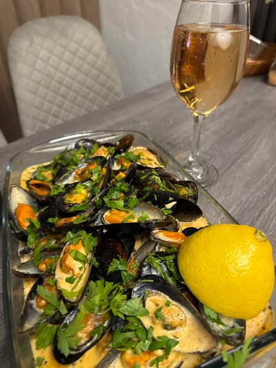 A close-up, high-angle shot captures a delicious meal of mussels in a creamy sauce, generously garnished with chopped parsley. The mussels are served in a clear glass baking dish, and a bright yellow lemon rests on the edge of the dish. In the background, a tall stemmed glass filled with a golden-hued beverage, likely white wine or champagne, sits on a grey textured surface, possibly a table. A glimpse of a quilted-back chair in a neutral tone suggests a dining setting. The scene is set in Sustaš, Montenegro, indicating a local culinary experience.